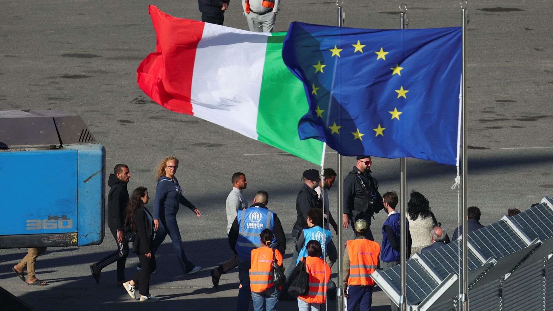 Migrants and security officials walk next to the EU and Italian flags as they disembark from the Italian navy ship Libra in Shengjin. /Florion Goga/Reuters