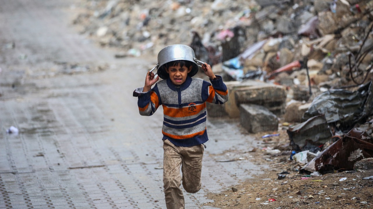 A displaced boy covers his head with a pan as he runs from the rain past building rubble at the Bureij refugee camp in the central Gaza Strip. /Eyad Baba/AFP