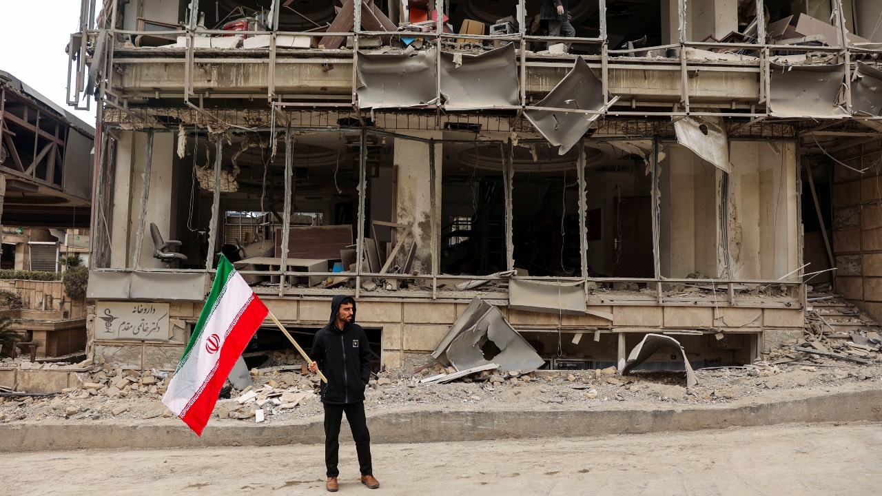 A man holds an Iranian flag following an Israeli/US strike on Gandhi Hotel Hospital. /Majid Asgaripour/WANA/Reuters