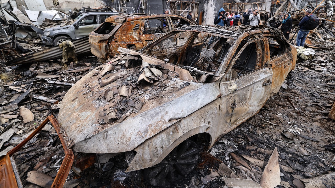 Iranian officials and journalists stand next to the wreckage of vehicles during the visit of a car service center in eastern Tehran that was hit by a missile strike. /Atta Kenare/AFP