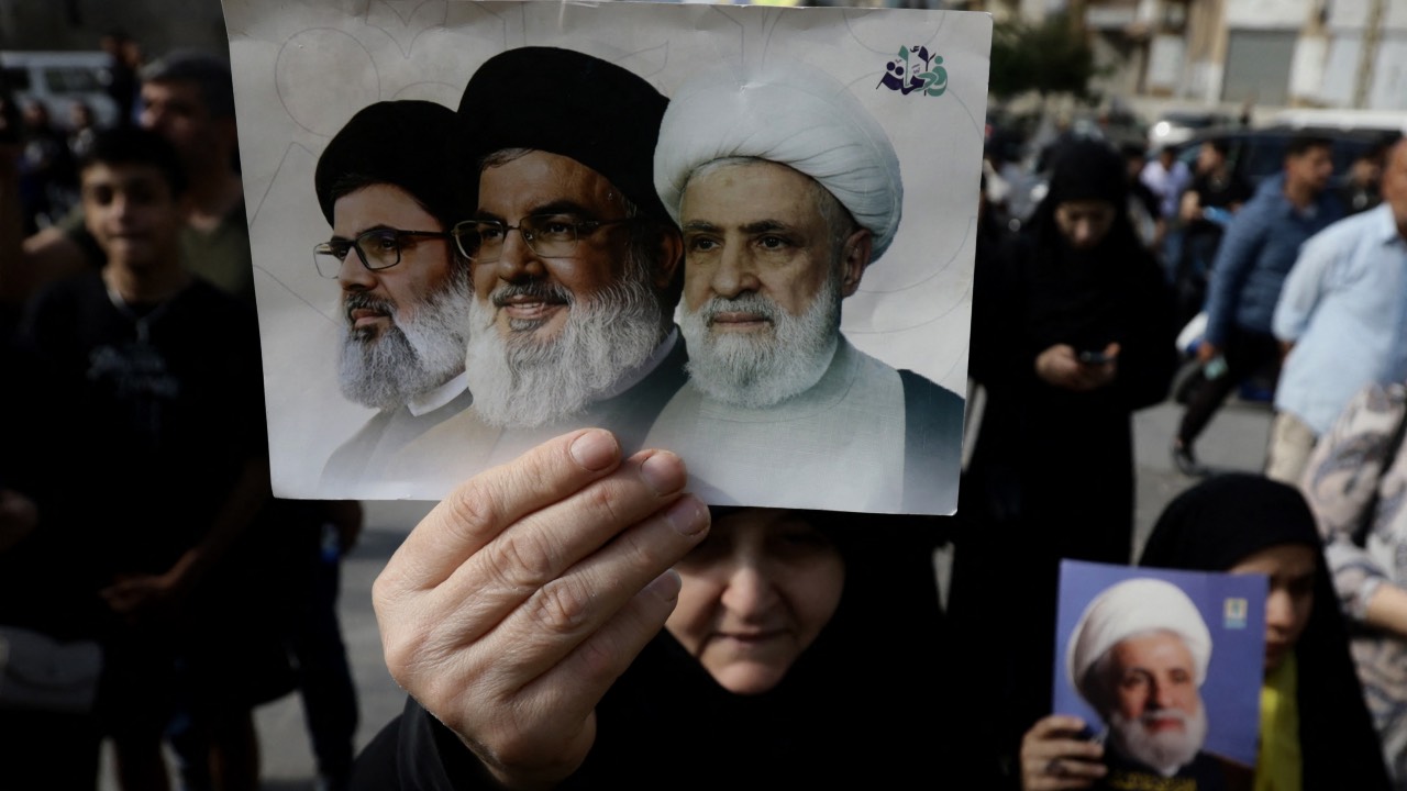 A Hezbollah party supporter holds a large postcard with the images of Lebanese Shiite Muslim Hezbollah leaders assassinated by Israel, Hashem Safieddine (L) and Hassan Nasrallah (C) and current leader Naim Qassem, during a solidarity rally against the United States and Israel, in Beirut’s southern suburbs. /Ibrahim Amro/AFP
