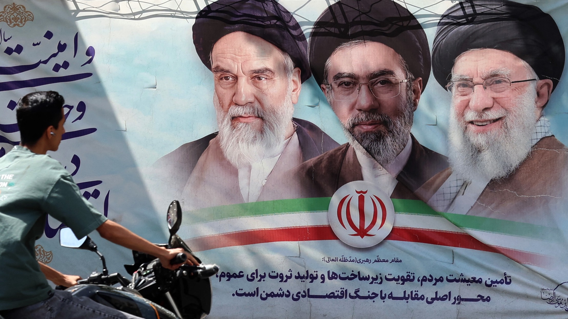 An Iranian man rides his motorbike past a billboard bearing the images of the late founder of the Islamic Revolution supreme leader Ayatollah Ruhollah Khomeini (L), the late Iranian supreme leader Ali Khamenei (R), and his son the current supreme leader Mojtaba Khamenei (C), along a street in Tehran. /AFP
