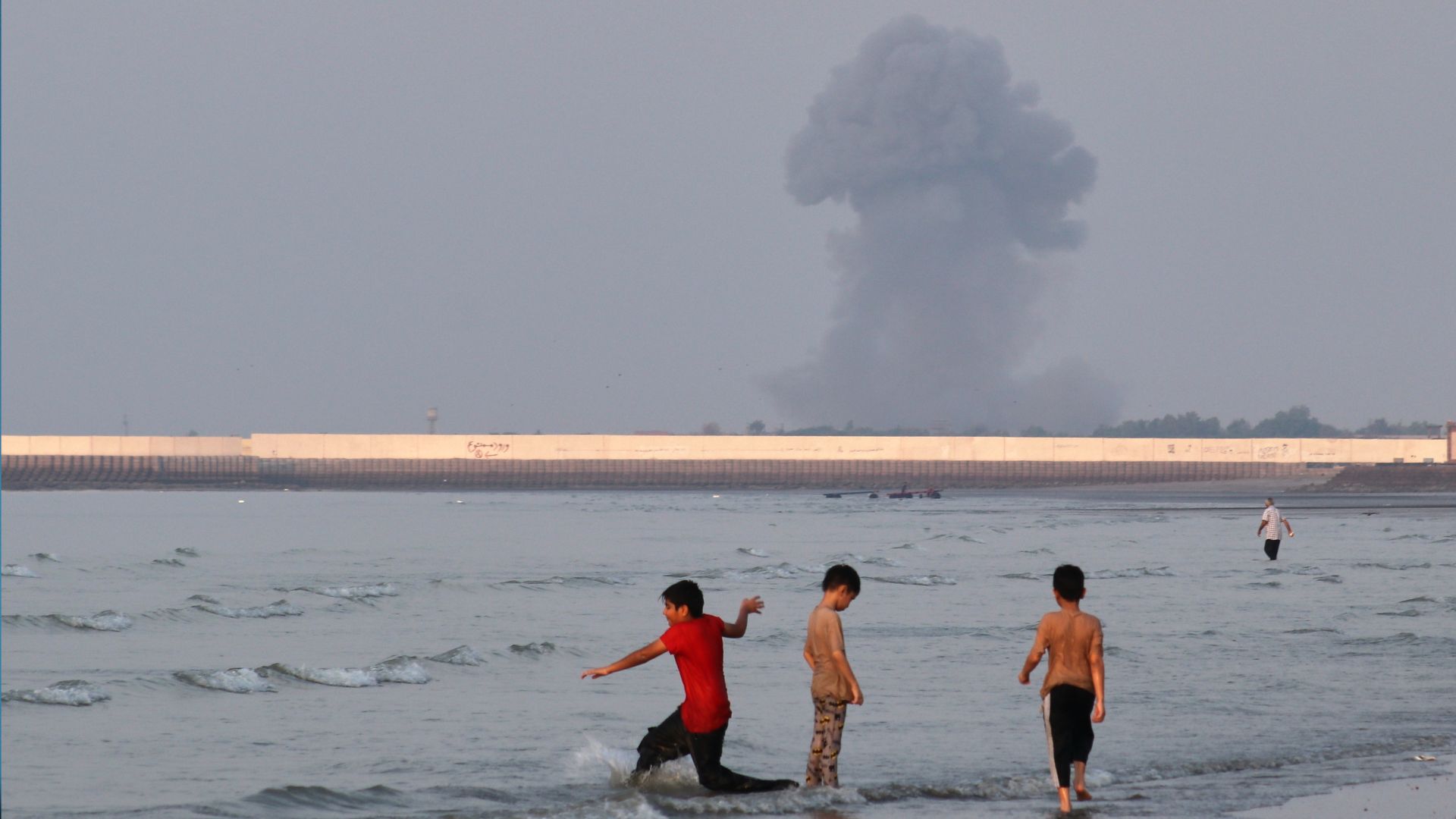Three boys play in the shallow waters of the Strait of Hormuz, as a plume of smoke rises from an explosion in the background, off Bandar Abbas, Iran, on July 13. /Razieh Poudat/ISNA via AP