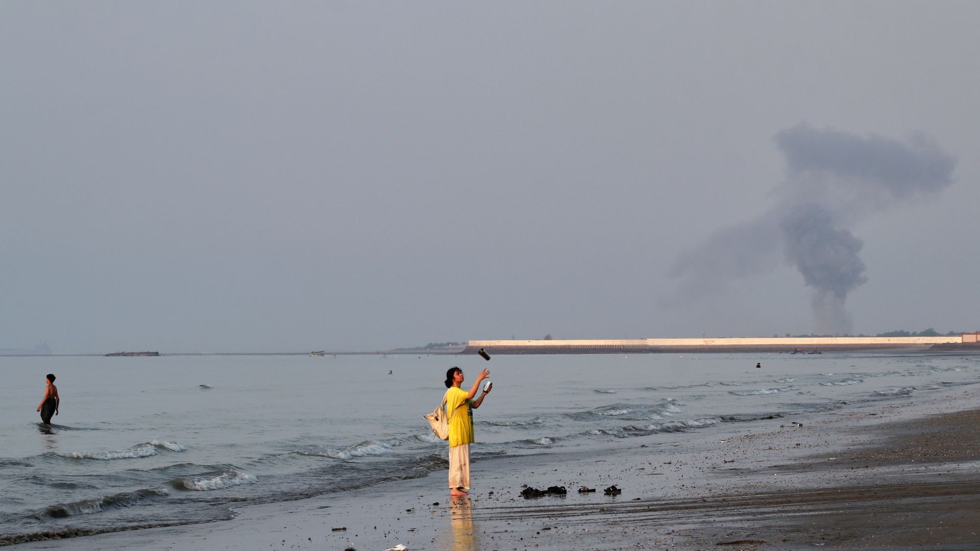 A woman stands at the water's edge along the Strait of Hormuz as a plume of smoke rises in the background following an explosion, off Bandar Abbas, Iran. /Razieh Poudat/ISNA via AP