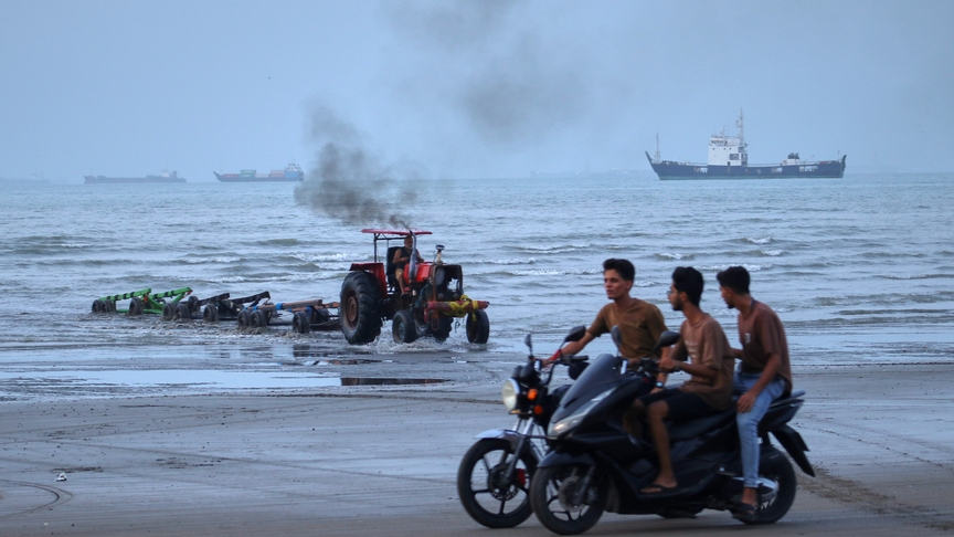 Young men ride motorcycles along the shore as a tractor pulls equipment from the water and commercial vessels appear anchored in the Strait of Hormuz off Bandar Abbas, Iran, Monday, July 27, 2026. /Razieh Poudat, ISNA via AP