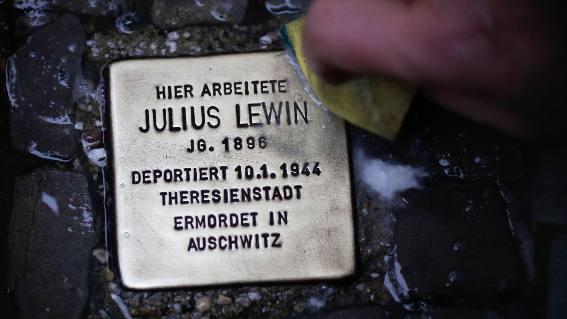 A polished 'stumbling stone' catches the light on a dark pavement. This stone commemorates the life of Julius Lewin, who died in Auschwitz. /AP