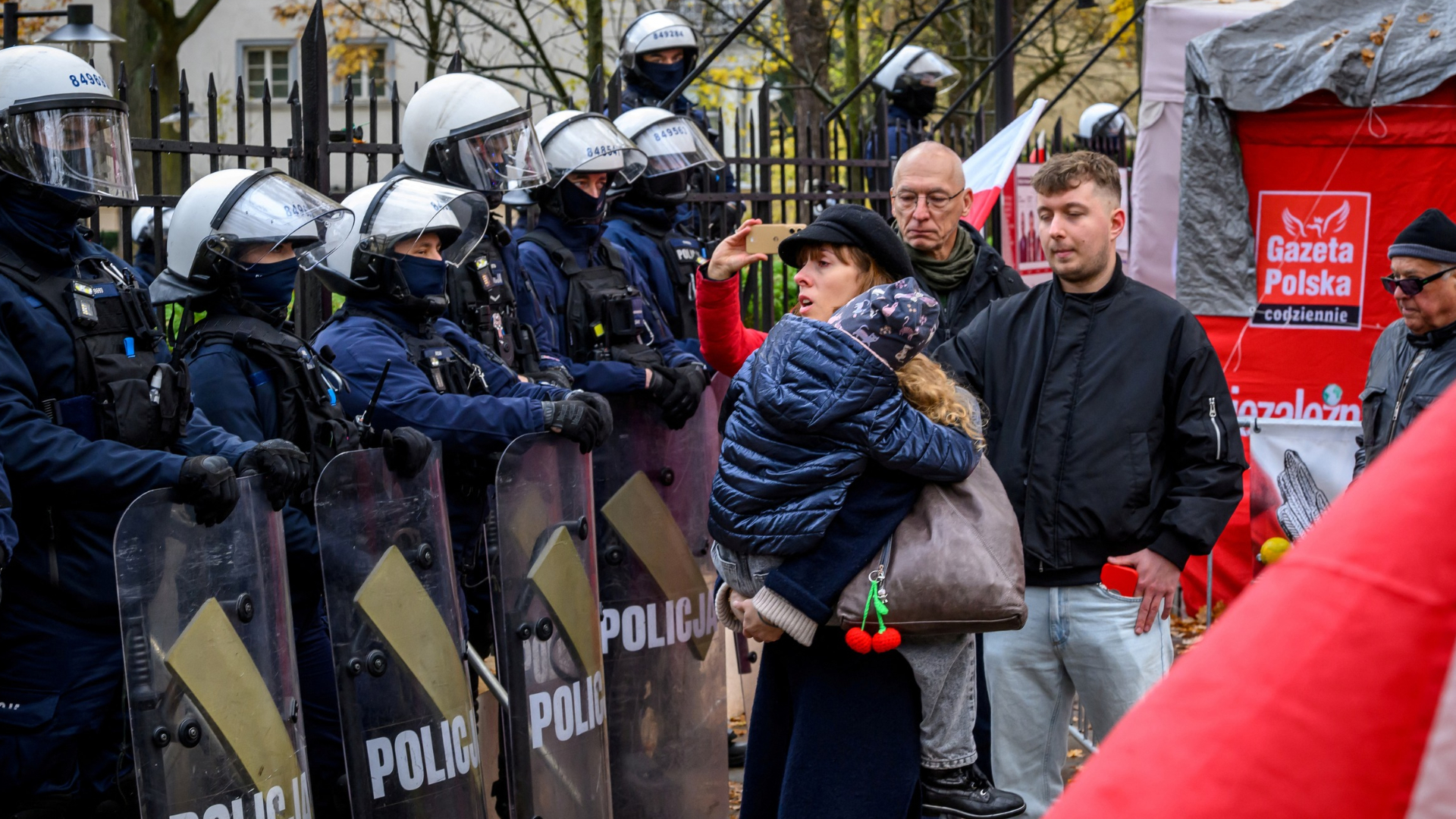 A woman carrying a child stands in front of police officers in riot gear as protesters gather outside the headquarters of Poland's Constitutional Court. Mladen ANTONOV / AFP. 31 October 2026.